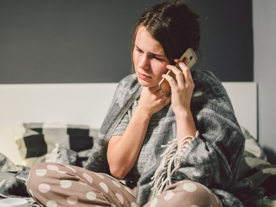 Eine Frau mit Schal sitzt auf dem Bett. Sie hat Halsschmerzen und hat ein Telefon in der Hand.