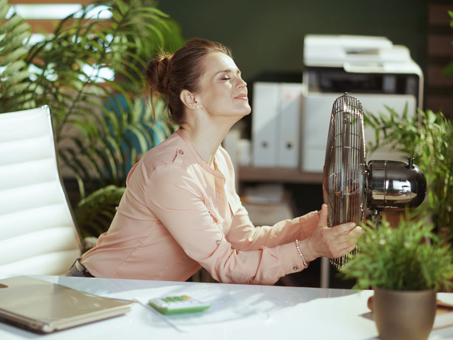 Eine schweissgebadete Frau sitzt an einem Ventilator im Büro.