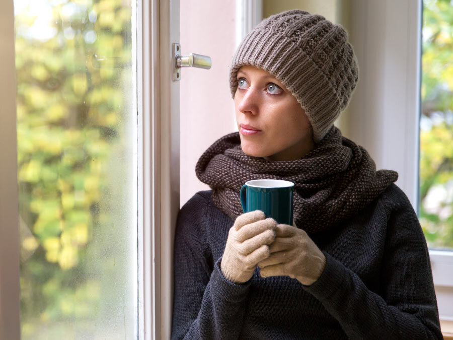 Eine Frau sitzt frierend mit Schal und Mütze am Fenster. In der Hand hält sie eine Tasse in der sich ein warmes Getränk befindet.
