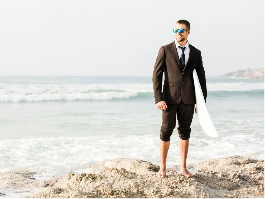 Ein Mann im Anzug steht am Strand. In der Hand hält er ein Surfbrett.