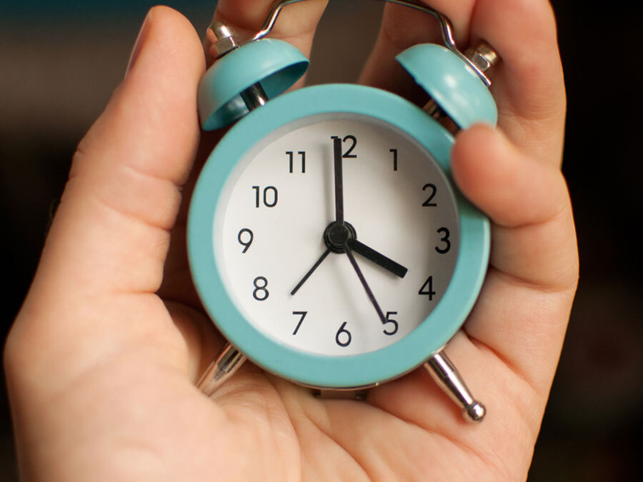 Eine Hand hält einen kleinen blauen Wecker, der auf vier Uhr anzeigt.