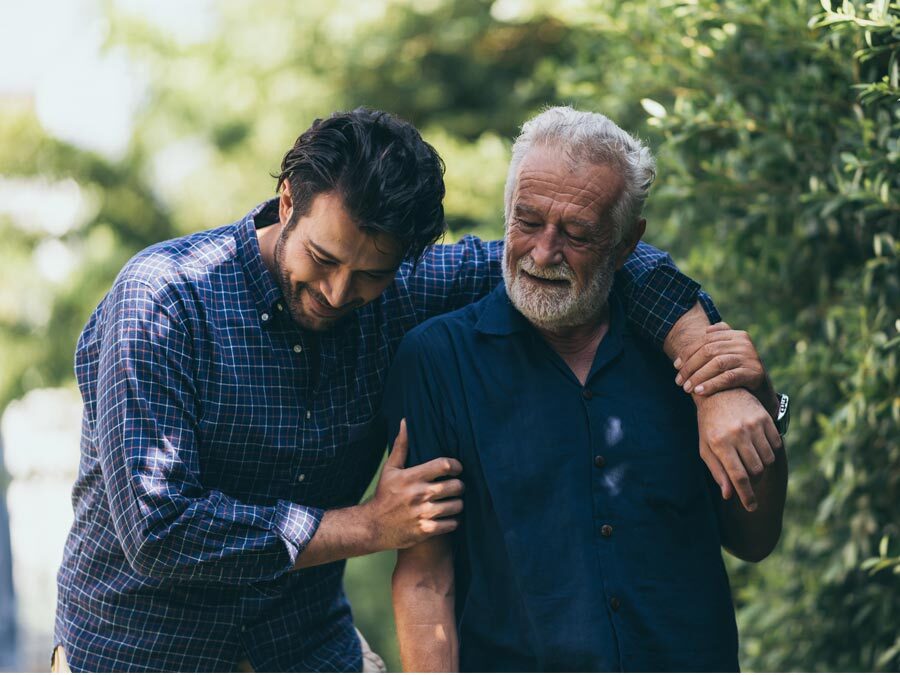 Ein älterer Herr mit grauen Haaren wird von einem jungen Mann, seinem Sohn, gestützt.