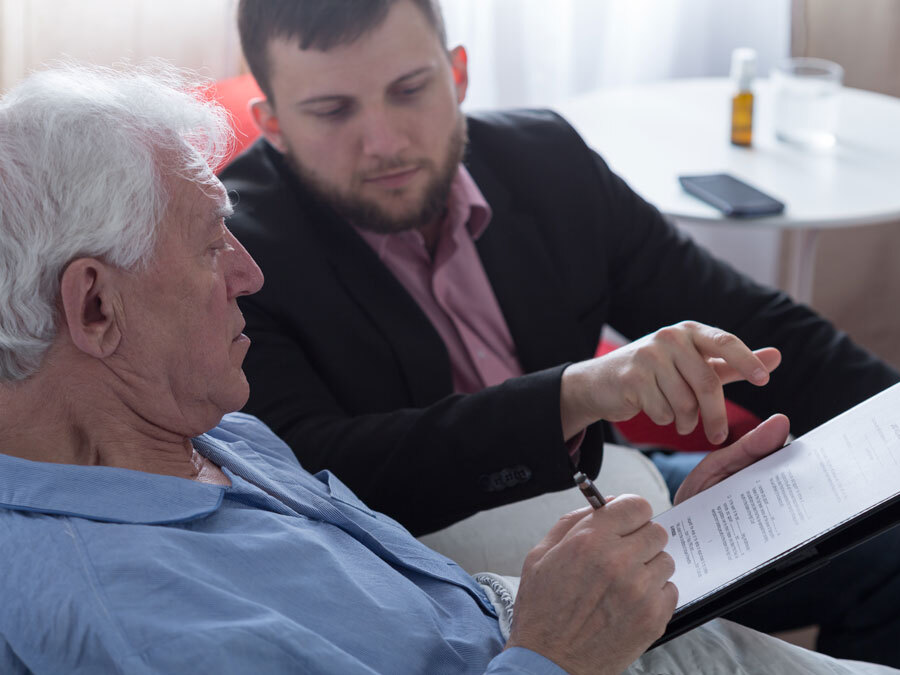 Am Krankenbett hält ein junger Mann einem alten Mann ein vorgeschriebenes Testament hin.