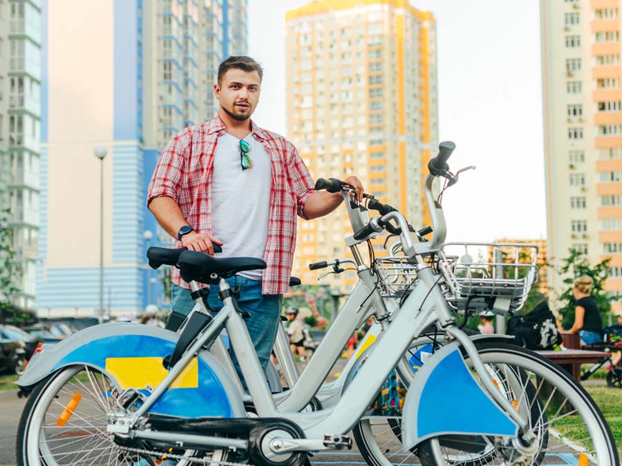 Ein junger Mann steht vor zwei hohen Gebäuden und hat ein Mietrad in der Hand.