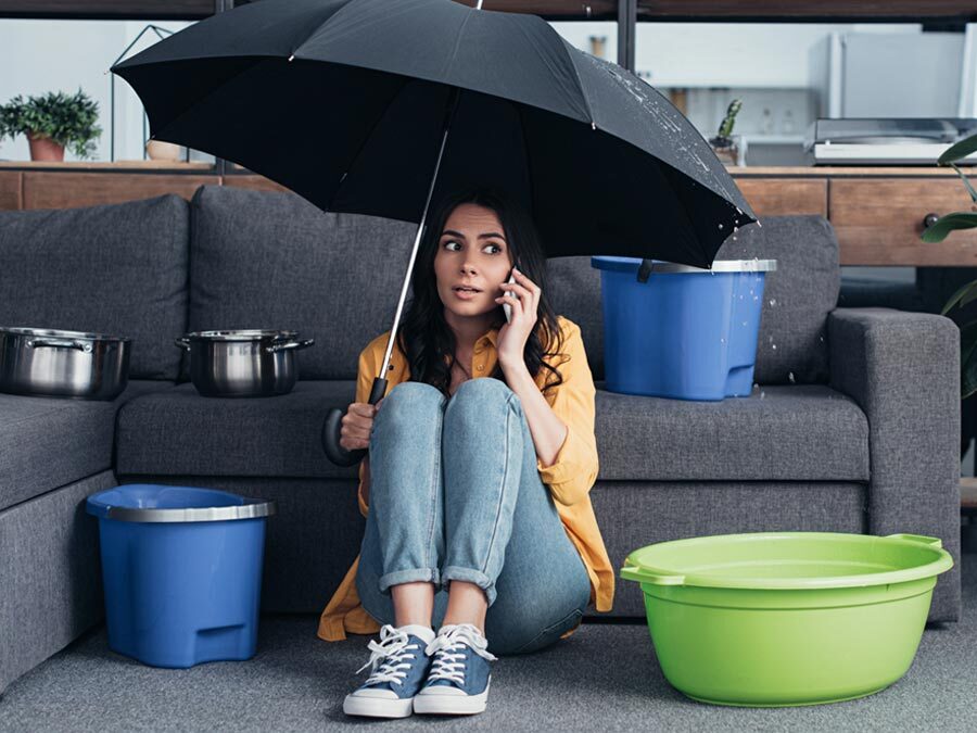 Eine Frau sitzt mit einem Regenschirm im Wohnzimmer. Neben ihr sind Schüsseln aufgestellt. Es regnet rein, denn sie hat einen Wasserschaden.