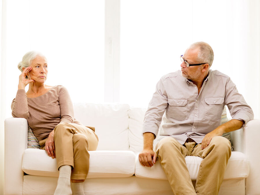 Eine Frau mit grauen Haaren und ein Mann im gleichen Alter sitzen auf einem Sofa und schauen sich verärgert an.