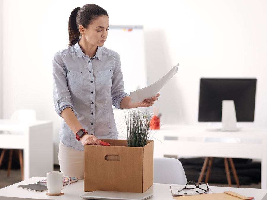 Eine Frau packt ihre Sachen auf dem Schreibtisch im Büro zusammen. In der Hand hält sie die Kündigung zum Arbeitsvertrag.