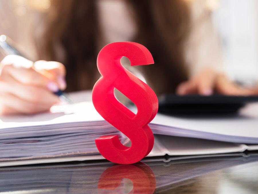 Wir sehen die Hand einer Frau, die Unterlagen ausfüllt. Im Vordergrund ein Paragraphenzeichen.