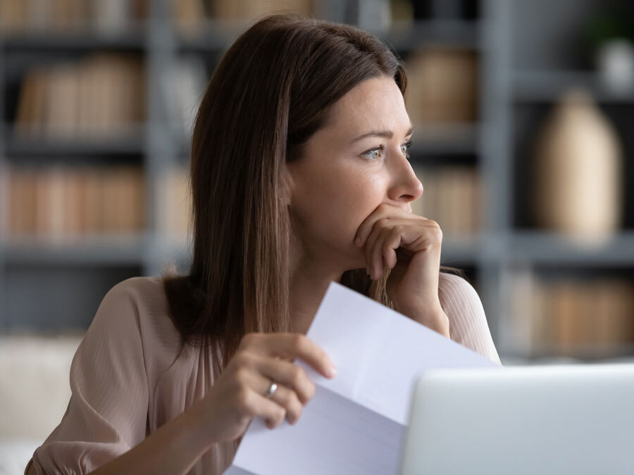 Eine Frau schaut nachdenklich aus dem Fenster. In der Hand hält sie das Schreiben eines Gläubigers. Wegen Erbschulden muss sie ein Nachlassinsolvenzverfahren einleiten.