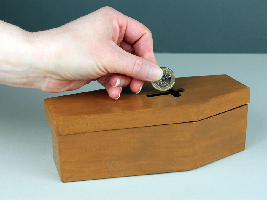Symbolisch legt eine Hand Geld in eine Spardose. Die Spardose ist wie ein Sarg geformt. Beerdigungskosten sind teuer.