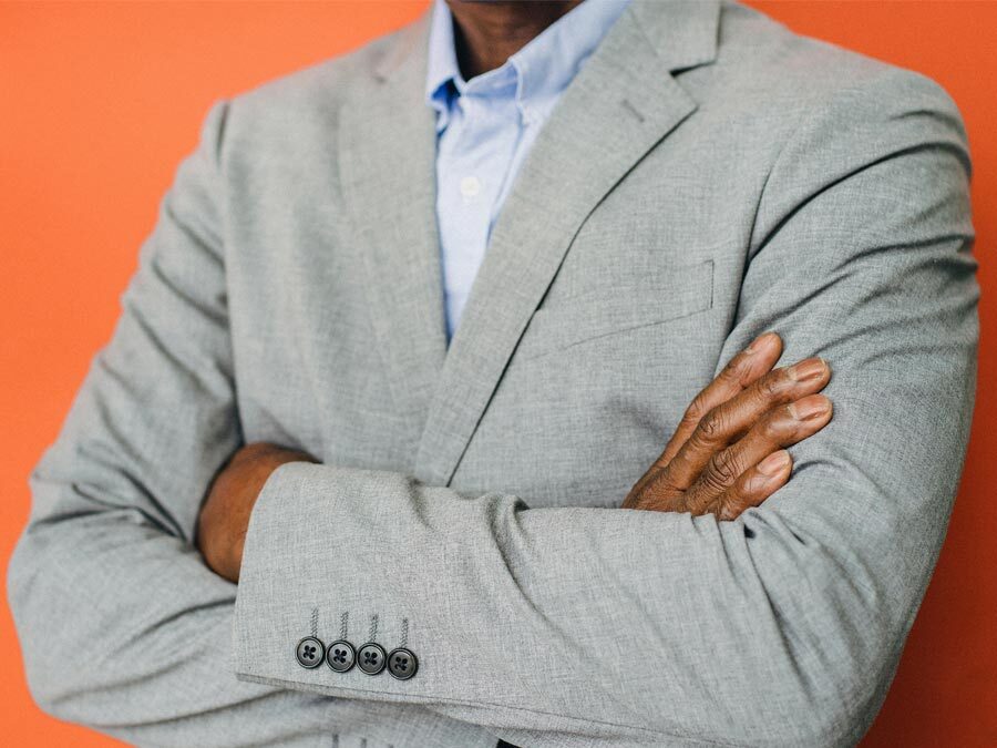 Ein Mann im grauen Anzug steht mit verschränkten Armen vor einer orange gestrichenen Wand.