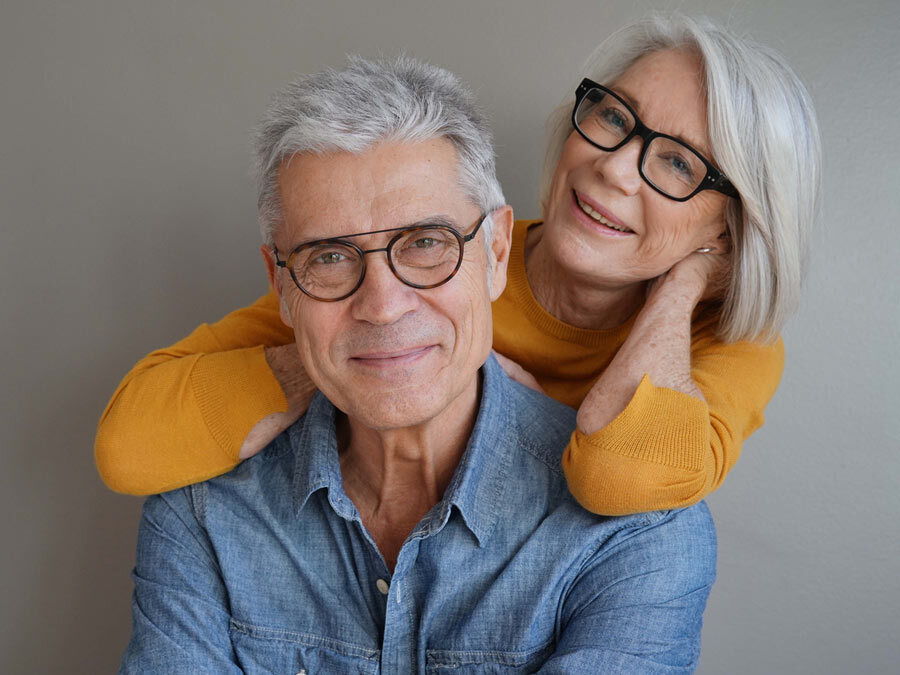 Eine Frau mit grauen Haaren lehnt sich an einen Mann mit grauen Haaren. Beide blicken in die Kamera.