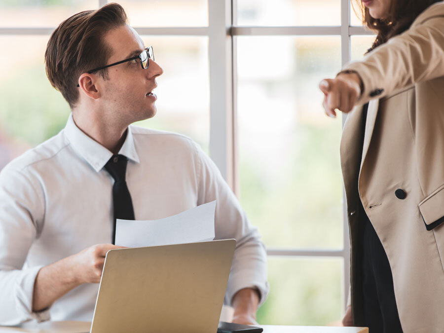 Ein Arbeitnehmer schaut entrüstet und fassungslos. Daneben sieht man seine Chefin, die ihn zururechtweist.