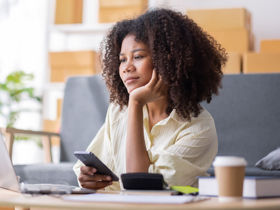 Eine Frau sitzt mit Smartphone vor dem Laptop. Hinter ihr stapeln sich Pakete.